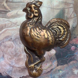 以下が含まれることがあります： テクスチャ加工されたボディと持ち上げられた尾を含む、詳細な特徴を持つブロンズ色の雄鶏の置物。雄鶏はテクスチャ加工されたベースの上に立っています。