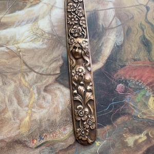 May include: A bronze-colored, rectangular decorative item with a floral and feminine face design. The item features detailed carvings of flowers and a woman's face, with flowing hair. The overall design is symmetrical and ornate.