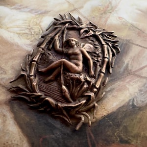 Könnte beinhalten: Bronze-farbener ovaler Anhänger mit einer nackten Figur, die in einem bambusartigen Rahmen sitzt. Die Figur ist detailliert dargestellt, und das Design hat eine antike Ästhetik. Der Anhänger ist vor einem Hintergrund mit gedämpften Farben platziert.