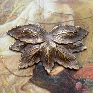 May include: A metallic decorative object shaped like a cluster of three leaves. The item features a dark bronze-toned finish with textured surfaces and polished gold-colored veins running through the center of each leaf, creating a layered appearance.