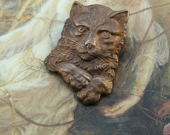 May include: A bronze-colored cat figurine with a detailed face and fur. The figurine is posed with its head turned to the side and its paw resting on its chin.