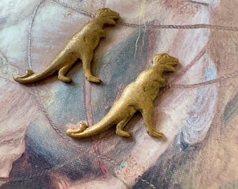 May include: Two gold-colored dinosaur figurines. The dinosaurs are in a standing position with their mouths open. The figurines are on a patterned background.
