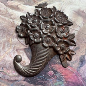 May include: A metal floral bouquet with a curled horn-shaped base. The bouquet is made of a cluster of stylised flowers with raised details. The horn is textured and has a rounded end.