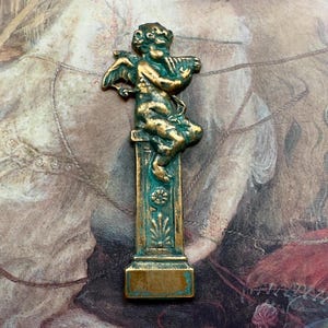 Puede incluir: Una figurita de querubín de metal verde y dorado tocando un instrumento musical, de pie sobre un pedestal con detalles intrincados.
