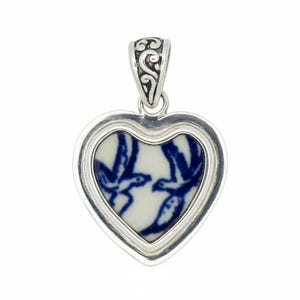 May include: Heart-shaped pendant with a silver frame and bail. The pendant features a white ceramic insert with a blue bird design. The bail has an ornate, scrolled design. The pendant is a jewelry item.