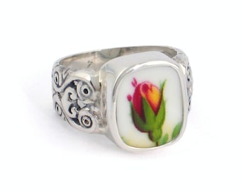 May include: A silver ring with a square-shaped porcelain cameo featuring a red and yellow rose with green leaves. The ring band has a decorative scroll design.
