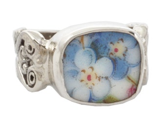 May include: A silver ring with a blue and white floral design. The ring has a square-shaped setting with a floral pattern on the band.