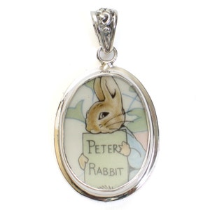 May include: A silver oval pendant with a white porcelain insert featuring a cartoon image of Peter Rabbit holding a sign that reads "Peter Rabbit".