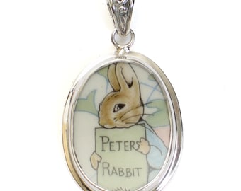May include: A silver oval pendant with a white porcelain insert featuring a cartoon image of Peter Rabbit holding a sign that reads "Peter Rabbit".