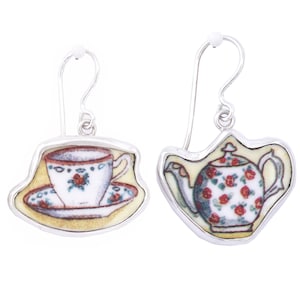 May include: Silver-tone earrings featuring a teacup and saucer design on one earring and a teapot design on the other. The teacup and teapot are white with red rose accents, set against a yellow background.