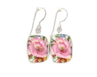 May include: A pair of drop earrings featuring rectangular ceramic pieces with rounded corners. Each piece displays a vibrant pink floral pattern with green leaves and brown accents, set in a silver-toned metal frame with silver-toned ear wires.