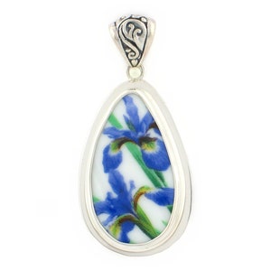 May include: A teardrop-shaped pendant featuring a white ceramic inlay decorated with blue iris flowers and green stems. The inlay is encased in a polished silver-toned metal frame with a decorative, engraved bail at the top for attachment.