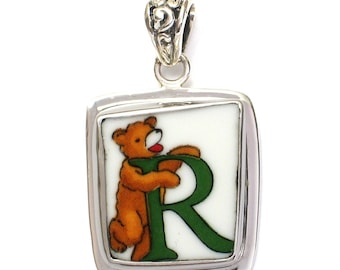 May include: A silver pendant with a white porcelain square featuring a cartoon bear holding a green letter R. The pendant has a decorative bail.