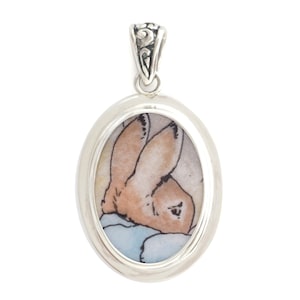 May include: A silver oval pendant with a detailed image of Peter Rabbit in a blue and white patterned fabric.
