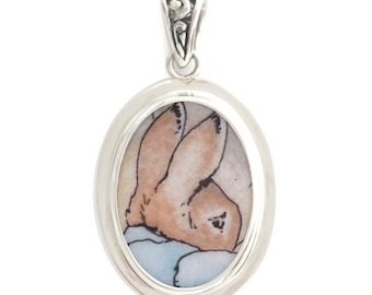 May include: A silver oval pendant with a detailed image of Peter Rabbit in a blue and white patterned fabric.