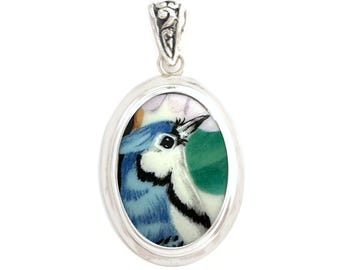 May include: An oval silver pendant featuring a detailed miniature painting of a blue jay bird. The bird has blue, white, and black plumage with a sharp beak. The pendant is framed in a polished silver setting with an ornate, decorative bail at the top.