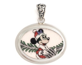 May include: A silver oval pendant with a white background featuring a cartoon image of Minnie Mouse wearing a Santa hat and holding a letter. The pendant has a silver bail.