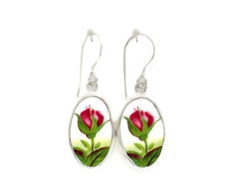 May include: A pair of silver drop earrings. Each earring features an oval-shaped porcelain insert with a red rose bud design. The rose bud is surrounded by green leaves and a yellow and green background. The earrings have a simple hook closure.