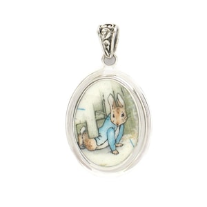 May include: An oval silver pendant featuring a detailed illustration of a brown rabbit wearing a blue jacket. The pendant has a polished silver frame and a decorative, ornate silver bail at the top for attachment to a chain or necklace cord.