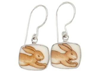 May include: A pair of silver drop earrings featuring square-shaped white ceramic tiles. Each tile displays a detailed illustration of a light brown rabbit in a running pose. The earrings are suspended from simple silver ear wires with a polished finish.
