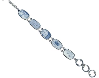 May include: A silver-toned link bracelet featuring five rectangular ceramic tiles decorated with blue and white illustrations of buildings, landscapes, and birds. The bracelet includes a toggle clasp and a series of circular rings at the end.