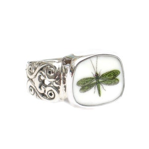 May include: A silver ring with an ornate band and a rectangular white porcelain top featuring a green dragonfly.