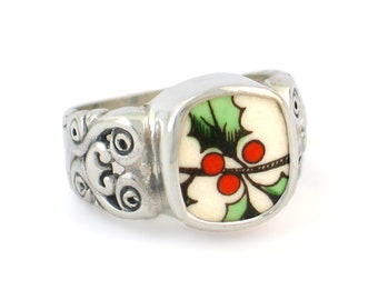 May include: A silver ring with a square-shaped, white ceramic inlay featuring a green holly sprig with red berries.