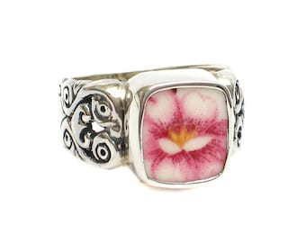 May include: A silver ring with a square-shaped pink and white flower design. The ring has a detailed, ornate band.