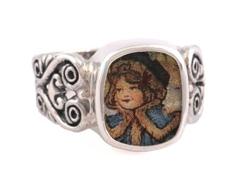May include: A silver ring with a square-shaped bezel featuring a vintage image of a girl wearing a blue coat and a hat. The ring band is decorated with intricate scrollwork.
