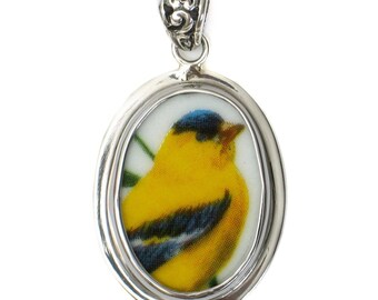 May include: Oval silver pendant featuring a yellow bird with blue and black accents. The bird is set against a white background with green details. The pendant has a decorative bail.