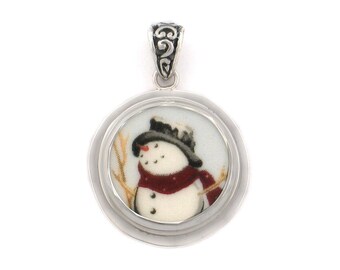 May include: A silver pendant featuring a round porcelain charm with a painted image of a snowman wearing a red scarf and black hat. The pendant has a silver bail.