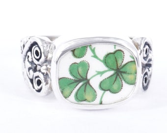 May include: A silver ring with a white oval-shaped setting featuring a green shamrock design. The band of the ring is decorated with a Celtic knot design.