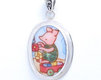 May include: An oval-shaped silver pendant featuring a colorful illustration of a small pink pig character wearing a green striped shirt. The pig is sitting next to a red toy wagon filled with blocks, set against a light blue background.
