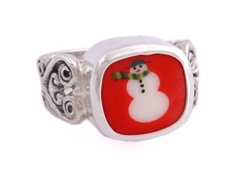 May include: A silver ring with a red square-shaped stone featuring a white snowman with a green scarf and black eyes. The ring has a decorative silver band.