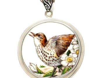 May include: A round silver pendant featuring a detailed hand-painted illustration of a brown and white speckled bird with its wings partially spread, perched near white flowers with yellow centers, set within a polished silver frame and bail.