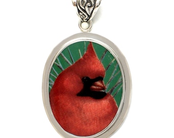 May include: An oval-shaped pendant featuring a vibrant red cardinal bird with a black face mask set against a green background with radiating lines. The pendant is encased in a polished silver-toned metal frame with an ornate, scrolled bail at the top.