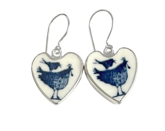 May include: A pair of heart-shaped drop earrings featuring a white background with a dark blue graphic print of a bird perched on top of a larger bird. The earrings are set in a silver-toned metal frame and include silver-toned ear wires.