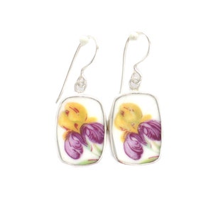 May include: A pair of drop earrings featuring rectangular ceramic pieces with rounded corners. Each piece displays a floral pattern with yellow and purple petals on a white background, set in a silver-toned metal frame with a curved hook wire.