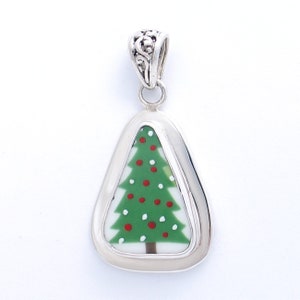 May include: A silver pendant with a white background and a green Christmas tree with red ornaments. The pendant is shaped like a triangle with rounded edges.