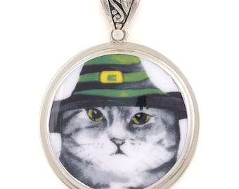 May include: A silver pendant with a round, white porcelain charm featuring a gray cat wearing a green and gold leprechaun hat.