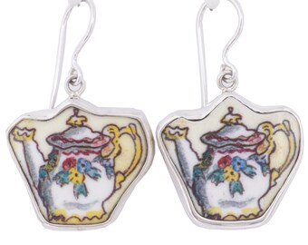 May include: A pair of silver dangle earrings featuring a white teapot with yellow, red, blue, and green floral details. The teapot is set in a silver frame.