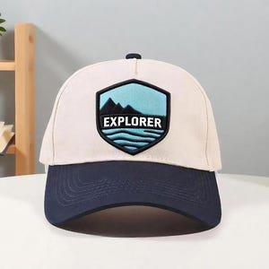 Puede incluir: Gorra de béisbol beige y azul marino con un parche en forma de escudo. El parche presenta un diseño de montaña y agua con la palabra "EXPLORER". La gorra tiene una visera curva.