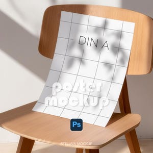 May include: A white rectangular poster mockup featuring a thin grey grid pattern and the text DIN A printed in black. The poster is displayed on a light-coloured wooden chair, with soft shadows of leaves cast across the surface of the paper.