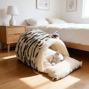 Puede incluir: Una acogedora cama para gatos con un estampado de rayas de tigre en blanco y negro. La cama tiene forma de túnel con un interior de color crema y un cojín a juego. Un gato durmiente descansa dentro de la cama, añadiendo a la escena acogedora.