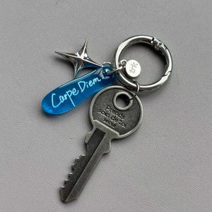 Puede incluir: Un llavero plateado con un dije en forma de llave y una etiqueta acrílica azul que dice "Carpe Diem". También se adjuntan un dije en forma de estrella y un dije circular con texto.