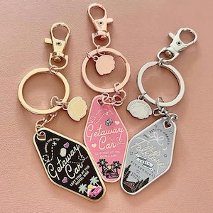 Könnte beinhalten: Eine Sammlung von Schlüsselanhängern mit verschiedenen Designs und Farben. Ein Schlüsselanhänger ist schwarz mit weißem Text und einer rosa Autografik. Ein anderer ist rosa mit weißem Text und einer Autografik. Der dritte ist silberfarben mit weißem Text und einer Hüttengrafik.