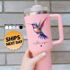 May include: A pink insulated tumbler with a handle, featuring a colorful hummingbird design. The tumbler has a clear lid and a straw. The name "Alicyn" is written below the hummingbird. The text "More Colors Available" and "SHIPS NEXT DAY" are also visible.