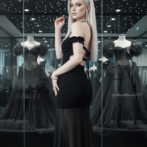 May include: A woman in a black dress with a sheer skirt stands in front of a glass display case. The dress has a cutout back and ruffled sleeves. Behind her are mannequins wearing black gowns with intricate details.