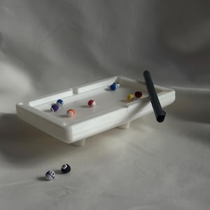 May include: A miniature white pool table with colorful billiard balls and a black cue stick. The table has a rectangular shape with rounded corners and small legs. Several balls are on the table, and two are off to the side.