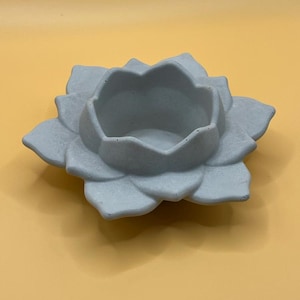 May include: A light gray, concrete lotus flower-shaped candle holder. The candle holder has a central well for a candle and multiple petals. The object is on a yellow background.